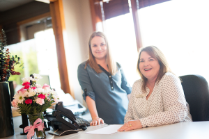 One woman standing and one woman sitting behind a desk with a phone and flowers in a vase